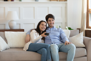 Overjoyed young caucasian couple sit relax on comfortable sofa in living room watching TV together, happy millennial man and woman rest on couch at home enjoy television program on leisure weekend