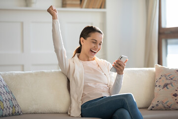 Overjoyed young Caucasian woman sit on sofa at home triumph get pleasant unexpected good message on...