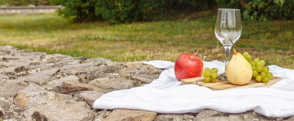 Picnic in the nature in the park. A glass of wine green grapes yellow pear red apple lilac on a wooden board on a white tablecloth in the grass in the garden on a stone path.