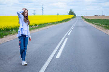 One caucasian beautiful girl in blue shirt and jeans, with  long brown hair  goes on empty road. Yellow background of blooming rapeseed field.  Rural landscape.