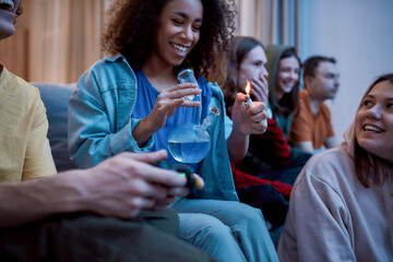 Friends smoking weed at home. Young afro american girl lighting marijuana in the glass bong, relaxing with friends on the sofa at home. Young people playing video games - obrazy, fototapety, plakaty