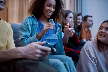 Fotobehang Friends smoking weed at home. Young afro american girl lighting marijuana in the glass bong, relaxing with friends on the sofa at home. Young people playing video games © Svitlana