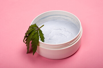 white round jar with a body scrub with a leaf of marijuana on a pink background close-up, cosmetics, skin care, beauty