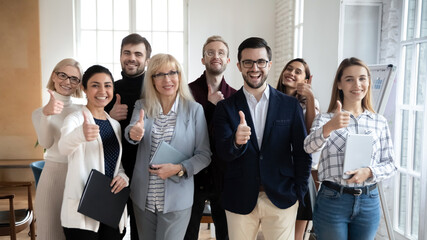 Excited group of diverse different ages businesspeople looking at camera, showing thumbs up gesture. Happy multiracial middle age and young employees evaluating seminar lecture, proposing services.