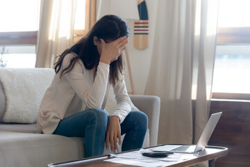 Unhappy young woman sit on sofa at home stressed about financial problems paying bills online,...