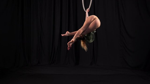 The flexible athletic circus gymnast performs acrobatic elements on the air ring. Circus performer spinning in a split on a background of black backstage scenes. young aerialist on aerial hoop.