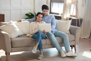 Happy young Caucasian couple sit relax on sofa in living room watching video on laptop together, smiling millennial man and woman rest on couch at home using computer, browsing wireless internet