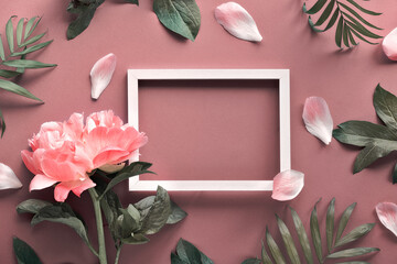 Creative flat lay, top view with light coral peony, petals and exotic palm leaves around white empty blank frame with copy-space on rosy brown or dark beige background.