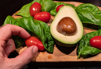 A wooden cutting board on a black background with an avocado in the center. All around, intense green basil leaves and bright red cherry tomatoes.  One hand touches a cherry tomato