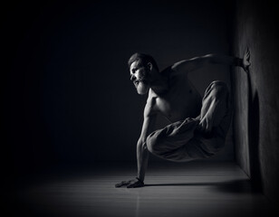 Aged man, naked torso, in brown pants is practicing yoga. Legs in lotus pose, setting one hand against wall, another one on floor