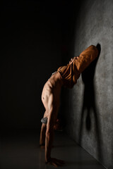 Aged male with naked torso, in brown pants is practicing yoga. Standing on hands, leaning against...