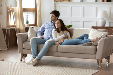 Happy dreamy young caucasian couple rest cuddle on couch in design living room thinking or...