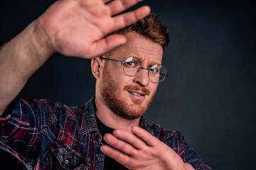 Take this ugly thing away from me. Displeased intense redhead mature guy in glasses and checkered shirt, turning away and protecting face with raised hands, seeing something disgusting over grey wall