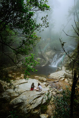 back of view two friends sitting near waterfall in forest