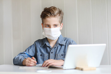 School boy with face mask watching online education classes at home. COVID-19 pandemic forces children online learning.