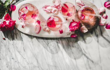 Summer refreshing cold beverage drink. Flat-lay of rose lemonade with ice cubes in glasses and fresh rose flower petals on oval serving board over grey marble table background, top view, copy space