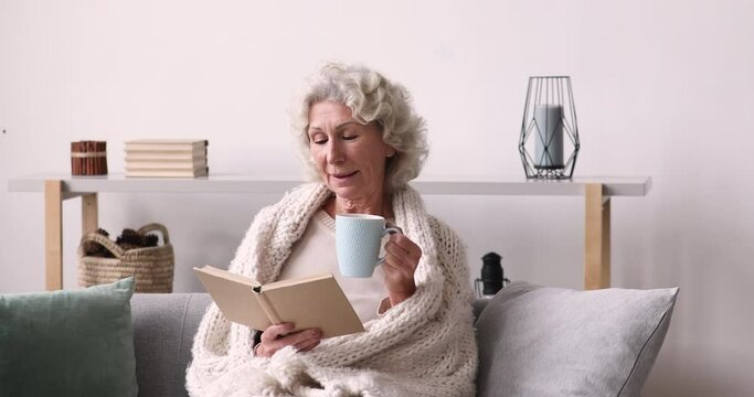 Dreamy relaxed middle aged mature woman reading favorite paper book novel bestseller, drinking hot coffee, sitting on sofa covered in plaid, spending cozy weekend at home during cold winter season.