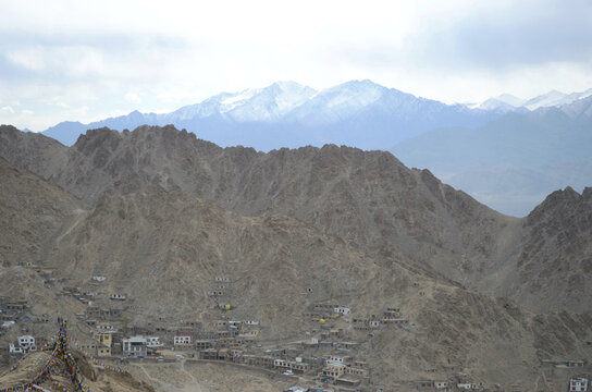 Prayer flags overlook a village in Ladakh, India, built into the slopes of a barren mountainside. Snow-capped mountains are in the distance, part of the Western Himalayas. The sky is overcast.