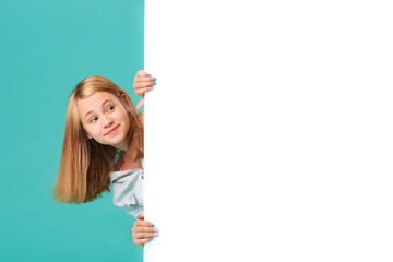 A smiling girl stands behind a white blank panel isolated against a turquoise background. Looks out...