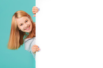 a laughing girl standing against a turquoise background holds an empty white Board in her hand and looks out from behind it