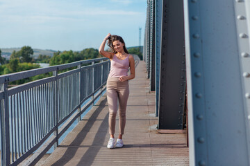 Obraz premium Pretty young woman posing on the old rusty transport bridge over the river during sunset.