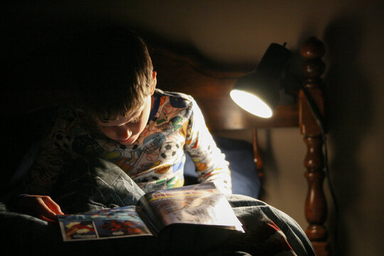 A young boy reads a comic book with a reading lamp in the dark while wearing pajamas.