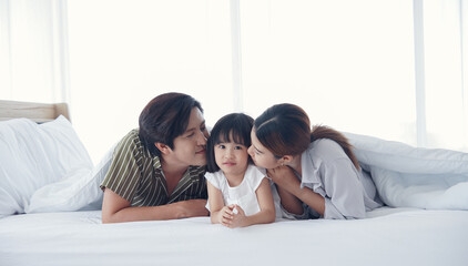 Asian Parents lying on bed under blanket and kissing their little daughter on both cheeks. family portrait