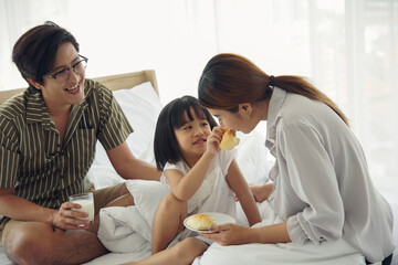 Obraz na płótnie Canvas A little cute daughter is giving bread sausage to her mother mouth. Asian family having breakfast on bed at bedroom