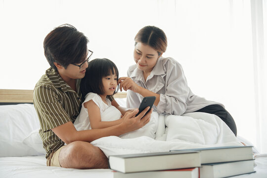 Happy asian young family spend time together. Dad hold mobile phone taking photo selfie with mom daughter who sitting under white blanket on the bed at bedroom