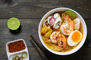 Spicy rice noodle with seafood and crispy wonton in white bowl on wooden table.