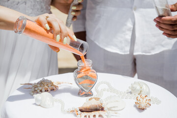 Obraz na płótnie Canvas Bride and groom pouring colorful different colored sands into the crystal vase close up during symbolic nautical decor destination wedding marriage ceremony on sandy beach in front of the ocean 