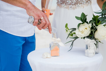 Bride and groom pouring colorful different colored sands into the crystal vase close up during symbolic nautical decor destination wedding marriage ceremony on sandy beach in front of the ocean 