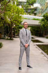 Full body shot of Hispanic bald bearded businessman wearing eyeglasses with nature in the city