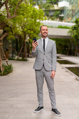 Full body shot of happy Hispanic bald bearded businessman thinking and using phone with nature in the city