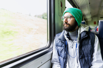 Stylish young man in glasses and headphones sits in a train car and looks out the window. Travel...