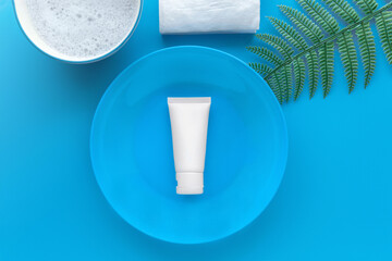 Top down view of organic gentle skincare product hyaluronic acid facial cleanser white tube for dry skin on a blue plate decoration with artificial fern leaves on plain blue isolated background.