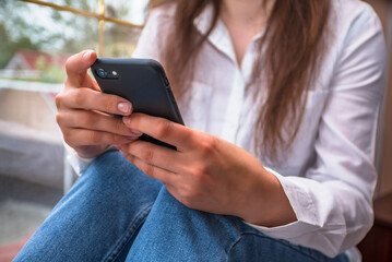 girl sitting at the window and holding a smartphone and watching social network close up hands and smartphone. Online shopping.