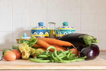 Vegetables on the cutting board in the kitchen with salt jars, spice jars and olive oil. Carrots, courgettes, aubergines, tomatoes, green beans.