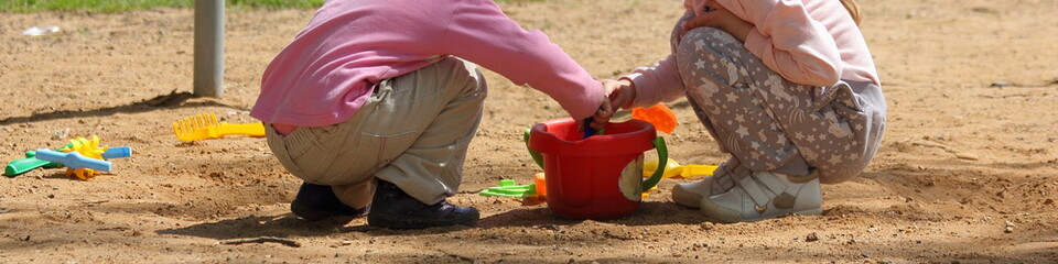 Two young children girls in demi season clothing play together in a big sandbox with a red bucket,...