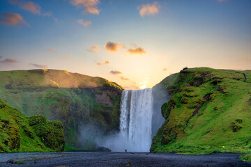 Men and women stand and raise their hands at the mighty Skogafoss Falls. In the morning, the sun rises from behind the mountains, with green grass all over the area. In southern Iceland