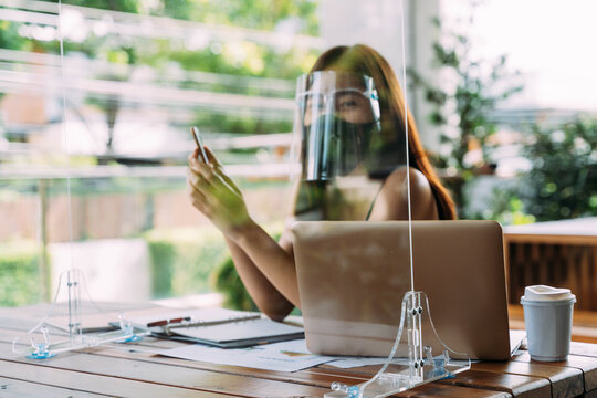 Young 20s Asian beautiful woman wearing a protective face shield and mask with partition in cafe restaurant. While using computer laptop and mobile phone outside. - Corona Virus prevention concept.