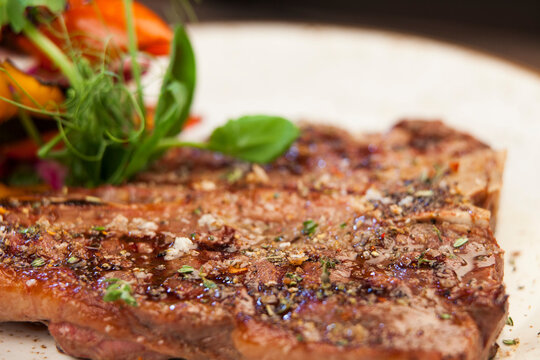 Close-up picture of different herbs and seasonings on a juicy steak on a white ceramic plate with some salad in the background