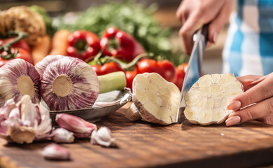 Detail of garlic head being cut to half with more fresh unpeeled garlic on the side and various veg in the background