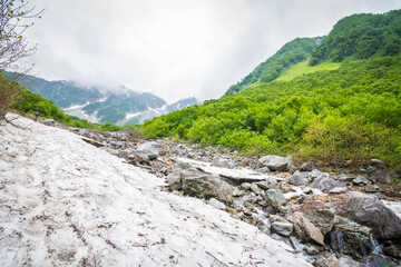 Fototapeta premium Unspecified hiker is climbing natural hiking route with green mountain rocky, cloudy sky and frozen ice from unmelted snow view. This is taken from Kamikochi National park, Matsumoto, Nakano Japan.