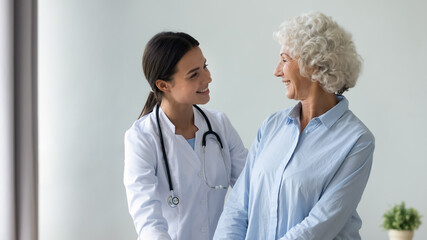 Caring young female caregiver in white coat smiling looking at old woman patient showing her...