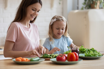 Caring young mother have fun chop vegetables preparing salad with cute little daughter at home, loving mom or nanny cooking breakfast together with small preschooler girl child in cozy kitchen