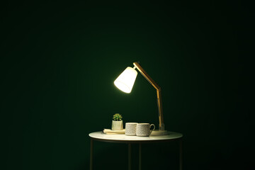 Glowing lamp, cups and houseplant on table in dark room
