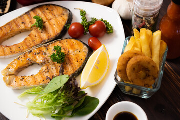 Delicious fillet cut salmon steak in white plate side dish with french fries and salad on dark wood background