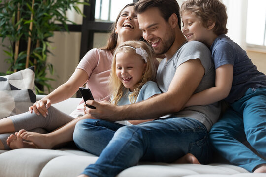 Happy young family with little children relax on sofa at home laugh watch funny video on smartphone together, smiling parents with small preschooler kids rest on couch have fun using modern cellphone