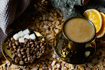 Aromatic dark coffee, with milk with bubbles on the surface in a dark gold Cup on a saucer, next to whole grains, peeled and dried oranges. Background of alder chips and wood snags in the background
